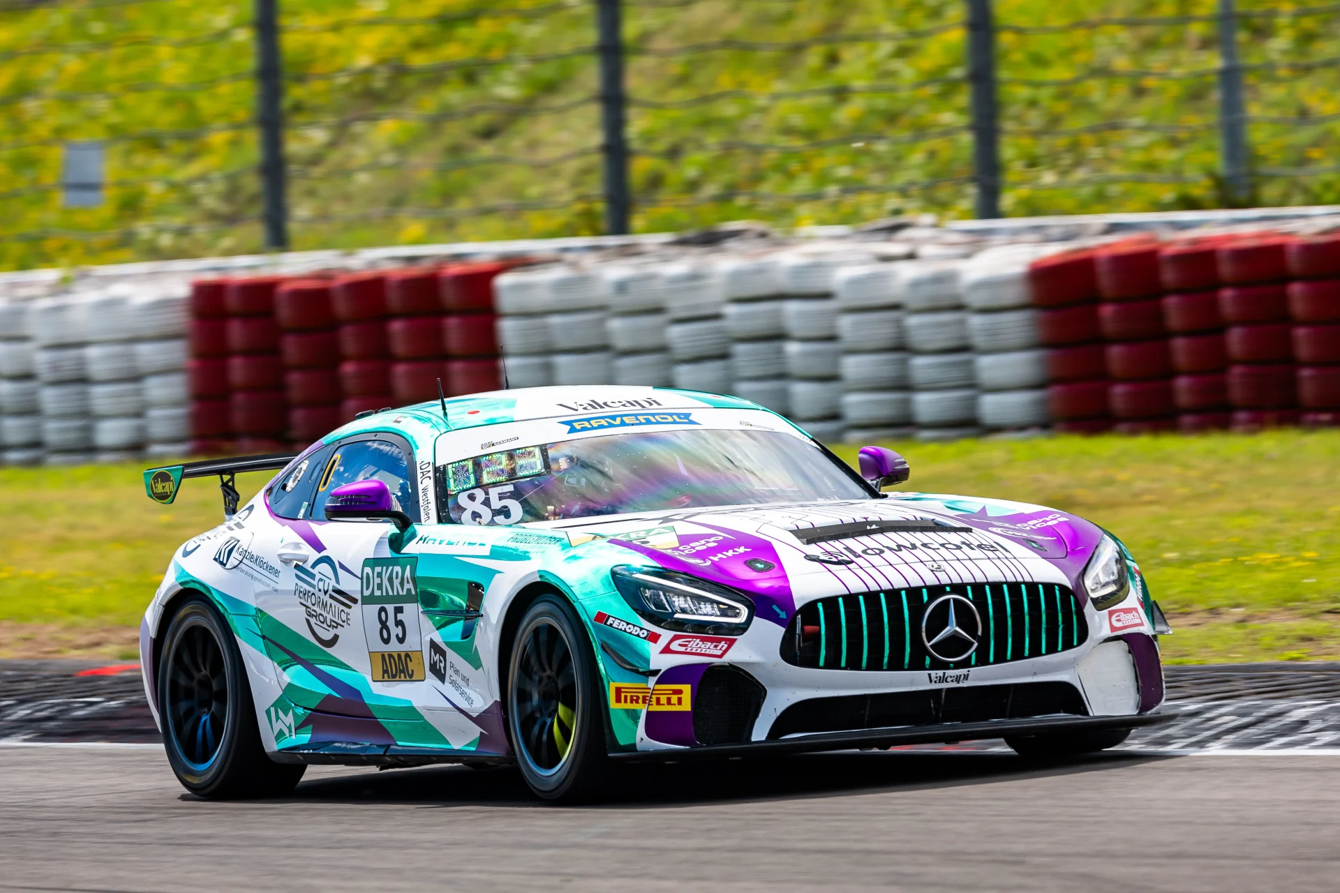 Ein farbenfroher Mercedes-AMG GT3-Rennwagen mit der Nummer 85 rast um eine Kurve mit verschwommenen Barrieren und grasbewachsenem Hintergrund und trägt auf seiner Karosserie verschiedene Sponsorenaufkleber.