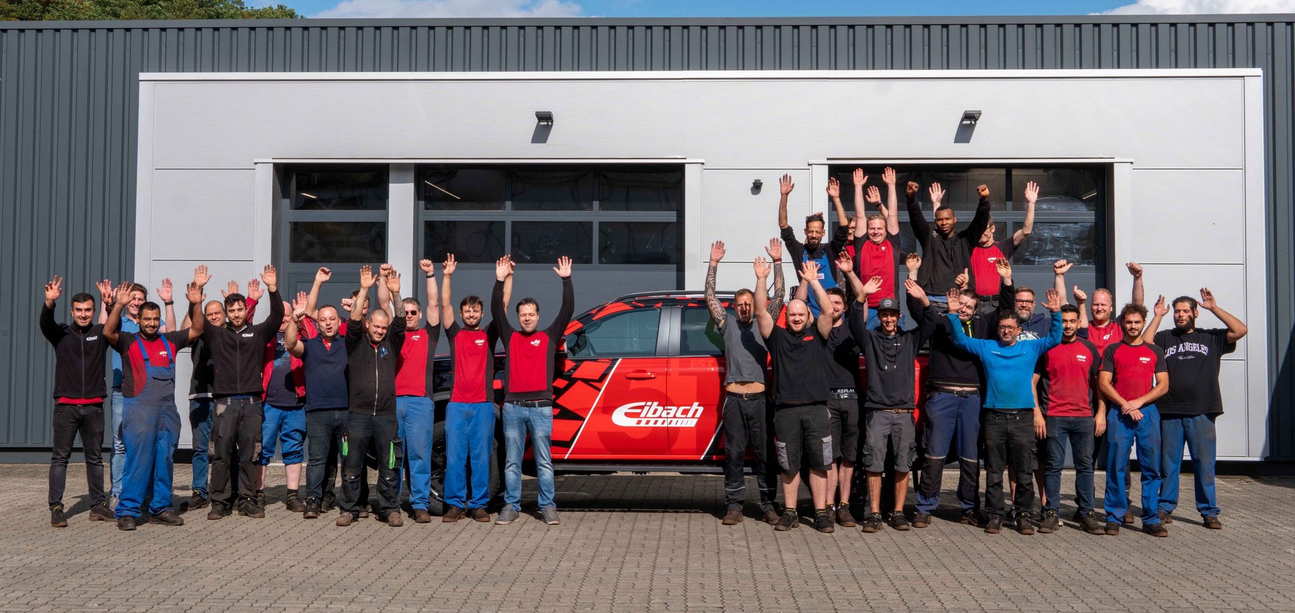 Eine Gruppe von etwa 30 Menschen steht vor einer grauen Industriehalle, streckt die Hände in die Höhe und lächelt. Dahinter parkt ein rotes Fahrzeug mit Eibach-Branding. Der Himmel ist klar und sonnig.