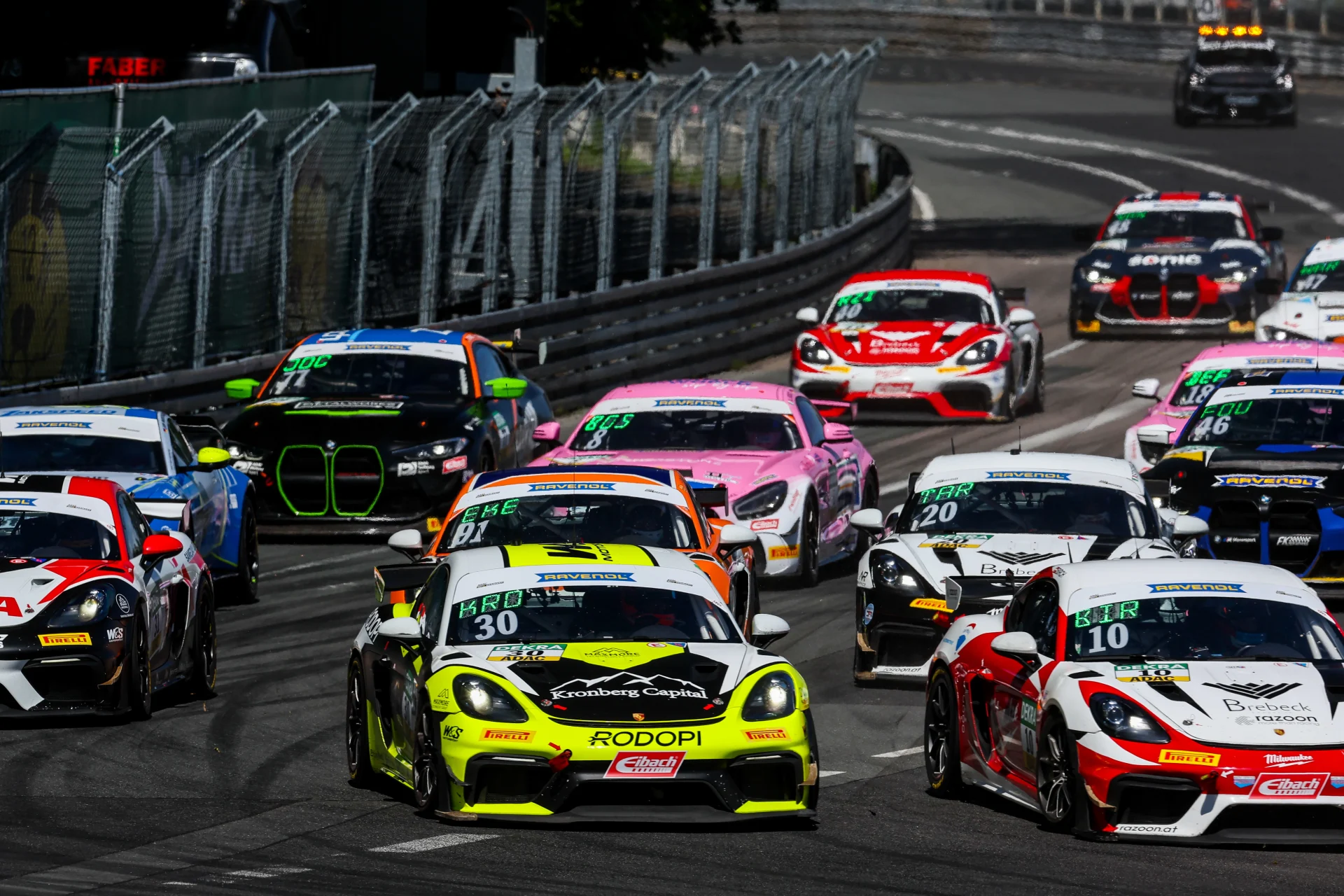 Während eines Motorsportwettbewerbs rast eine Gruppe bunter Rennwagen auf einem eingezäunten Stadtkurs um die Kurve. Die Autos stehen dicht gedrängt, und die Fahrer meistern die enge Kurve.