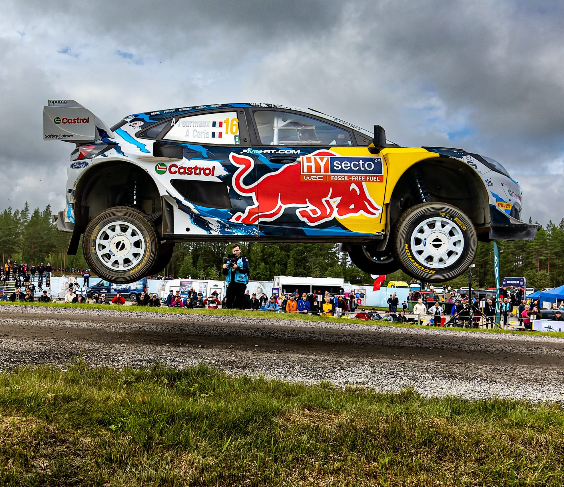 Ein Rallyeauto mit Red Bull-Branding springt während eines Rennens von einer unbefestigten Straße, mit Zuschauern und Bäumen im Hintergrund unter einem bewölkten Himmel.