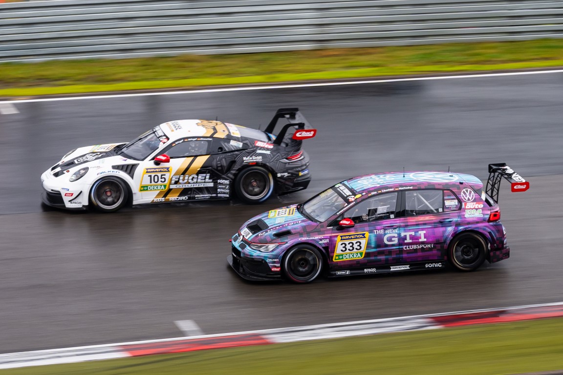 Zwei Rennwagen rasen Seite an Seite über eine nasse Rennstrecke, ein weißer Porsche mit der Nummer 105 und ein bunter VW mit der Nummer 333, mit Bewegungsunschärfe und Leitplanken im Hintergrund.