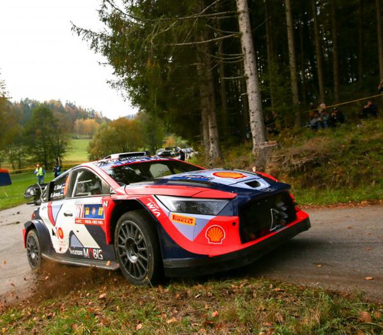 Ein Rallyeauto mit rot-blau-weißer Lackierung rast auf einer schmalen Straße um eine Kurve und wirbelt dabei Staub und Gras auf. Im Hintergrund sind Bäume und Zuschauer zu sehen.